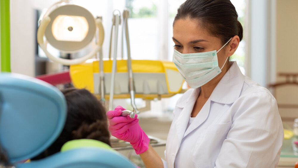 Treating caries. Serious woman doctor in pink aseptic gloves looking at a young female patient while holding a dental handpiece