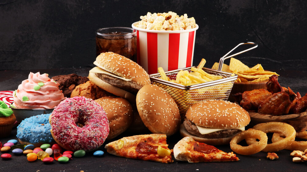 Hochverarbeitete Lebensmittel wie Burger, Doughnuts und Cup Cakes sind bunt aufgetürmt vor schwarzem Hintergrund. Hochverarbeitete Lebensmittel wie Burger, Doughnuts und Cup Cakes sind bunt aufgetürmt vor schwarzem Hintergrund.