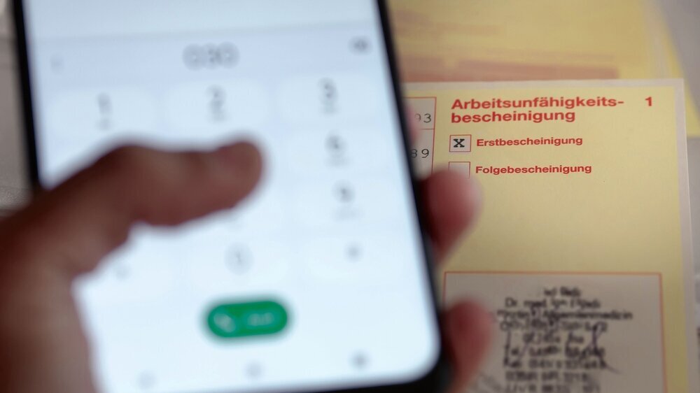 Telefonische Verlängerung der Krankmeldung: Hand wählt Nummer auf Smartphone und Arbeitsunfähigkeitsbescheinigung