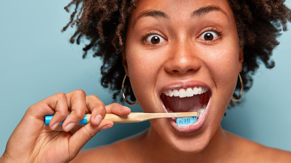 Glad mirthful woman with dark healthy skin, satisfied with new toothpaste, cleans teeth, opens mouth widely, has happy expression, curly hair, models over blue background. Close up portrait. Zähneputzen