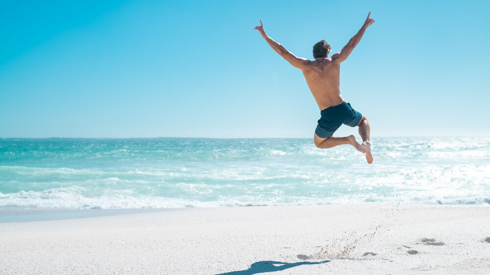 Rückansicht eines jungen Mannes, der mit ausgestreckten Armen am Strand freudig in die Luft springt.