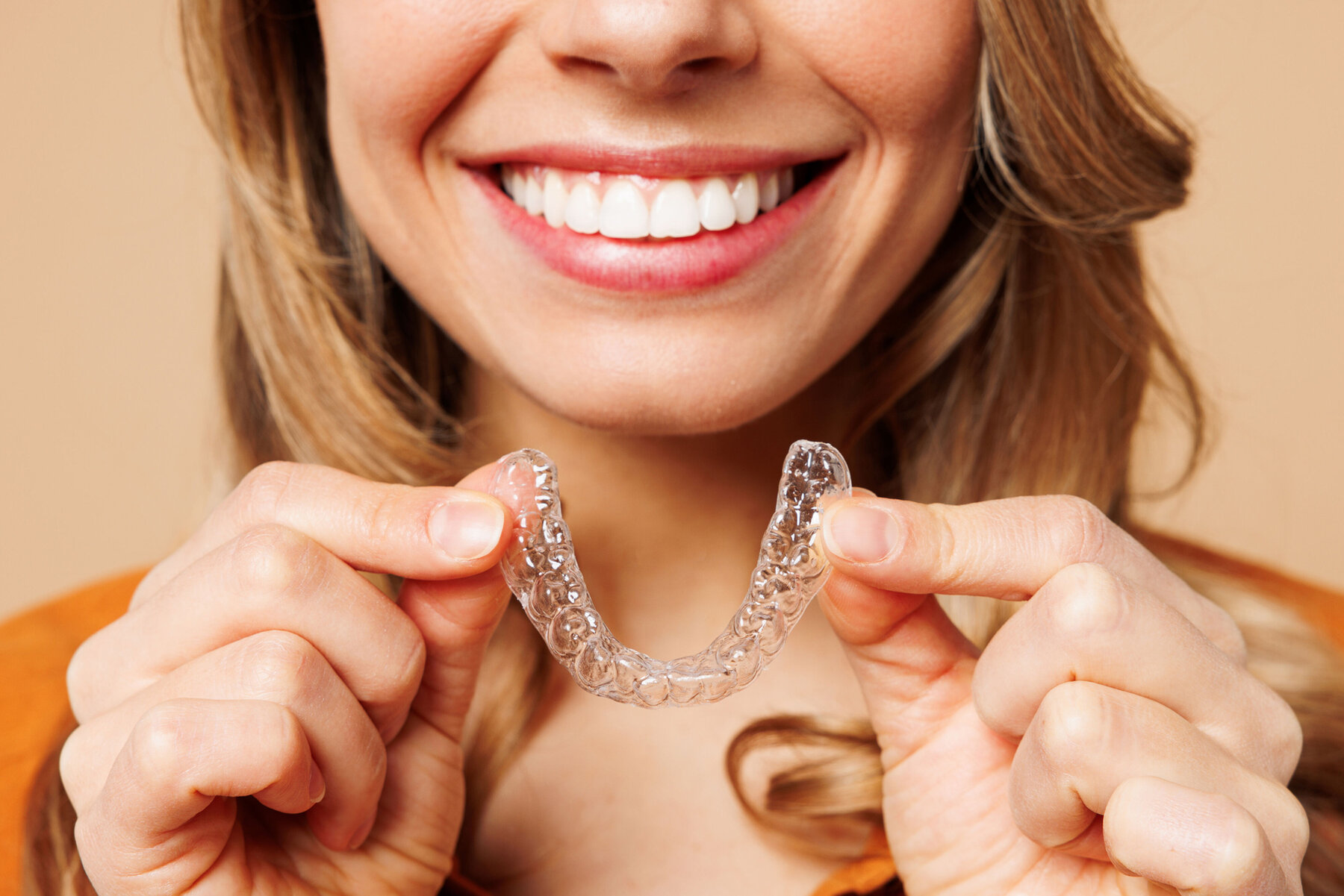 589663-flexible-1900.jpg Close up cropped young woman wear orange shirt casual clothes hold in hand invisible transparent aligners, invisalign bracer smile isolated on plain pastel light beige background. Lifestyle concept